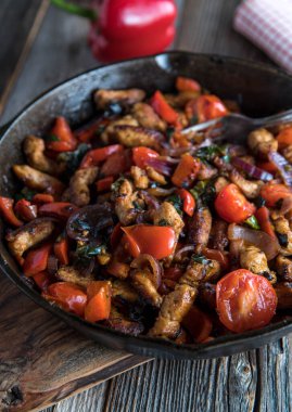 Healthy pan dish for low car diet with fried turkey breast, tomatoes, bell peppers, onions, garlic and herbs. Served in a rustic cast iron pan isolated on wooden table