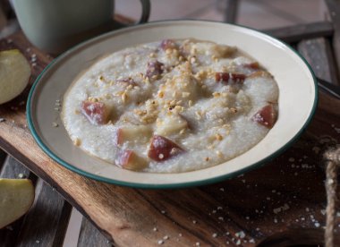 Rural breakfast with fresh cooked millet porridge, sauteed apples, roasted hazelnuts and maple syrup. Served in a old fashioned enamel bowl on wooden background
