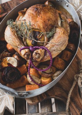 Roast whole chicken with root vegetables in a rustic roasting pan on wooden background