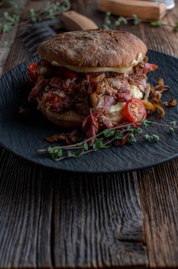 Pulled pork burger. Topped with roasted onions, cheese, pickles and tomatoes. Served isolated on a dark plate on old fashioned and wooden background with copy space