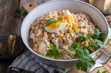 Homemade fresh cooked tuna salad with mayonnaise, boiled eggs, onions and corn. Served in a rustic bowl with poppy seed buns on wooden table background