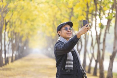 Süveter giyen Asyalı adam, sonbahar mevsiminde sarı ginkgo ağaçlarıyla parkta selfie çekmek için cep telefonu kullanıyor.