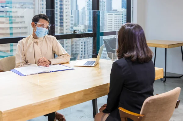 LGBTQ manager from HR department wearing facial mask is interviewing new applicant and looking at her resume and profile through the partition for social distancing and new normal policy