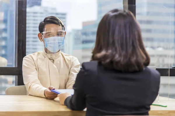 LGBTQ Manager from HR department wearing facial mask is interviewing new applicant who is handing her resume and profile through the partition for social distancing, transaction and new normal