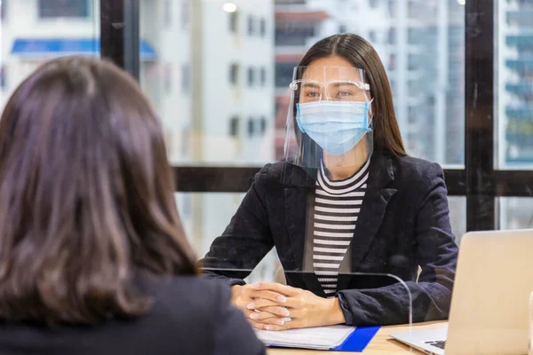 Manager from HR department wearing facial mask is interviewing new applicant and reading her resume and profile through the partition for social distancing and new normal