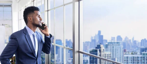 African American businessman with formal suit using mobile phone while looking out the window to the cityscape with skyscraper view for vision, dream and successful business and investment planning