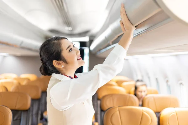 Asian female flight attendant closing the overhead luggage compartment for carry on baggage after passengers are seated and prepare to take off