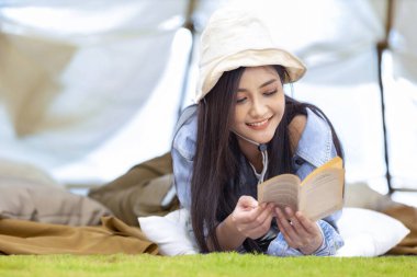 Asyalı kadın yaz mevsiminde macera ve aktif seyahat için ulusal parkta kamp yaparken çadırında kitap okuyor.