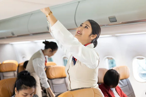 Asian female flight attendant closing overhead luggage compartment lid for carry on baggage after passengers are seated and prepare to take off