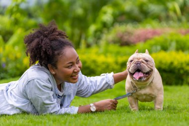 Afro-Amerikan kadın, parkta sabah egzersizi yaptıktan sonra çimlerin üzerinde uzanırken Fransız bulldog köpeğiyle oynuyor.