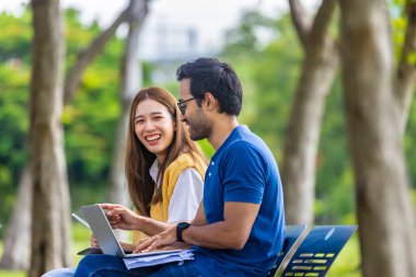 Genç Asyalı ve Hintli öğrenci hafta sonu boyunca bankta oturup ders çalışırken üniversite kampüsünde dinlenmenin keyfini çıkarıyorlar.