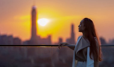 Woman looking and enjoying the sunset view from balcony with the sun setting behind skyscraper in busy urban downtown with loneliness for solitude, loneliness and dreaming of freedom lifestyle