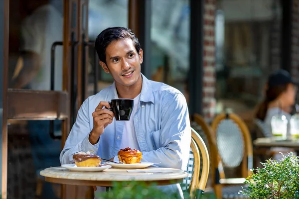 Asian man sipping a hot espresso coffee while sitting outside the european style cafe bistro enjoying slow life with morning vibe at the city square with the sweet pastry