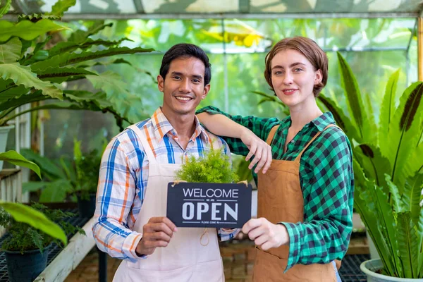 Team of diversity garden workers holding opening sign to welcome customer to their tropical nursery plant center full of exotic fern species