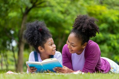African American mother is teaching her young daughter to read while lying down after having a summer picnic in the public park for education and happiness