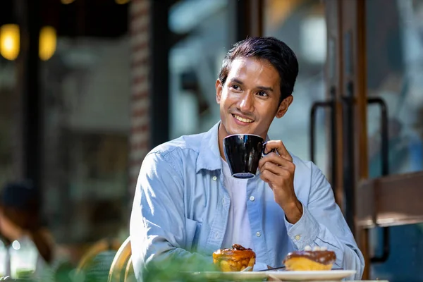 Asian man sipping hot espresso coffee while sitting outside the european style cafe bistro enjoying slow life with morning vibe at the city square with sweet pastry
