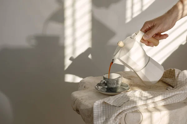 İtalyan kahve makinesinden ahşap saplı kahveyi koyu ahşap saplı gri espresso bardağına dökerek kremsi tonlarda keten çay havlusuyla süsleyerek güneş ışığında beyaz duvara yaslanarak güzel gölgeler yaratıyor. 