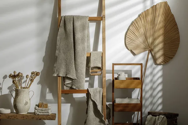 Organik el yapımı sabun, ahşap merdivende asılı keten havlular, bej sabun dağıtıcısı, deri yüzen vücut kayışı ve desenli vazoda kurumuş haşhaş çiçekleri. Sıfır atık banyo aksesuarı