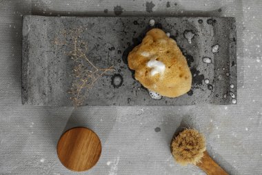 Gri beton tepside sabun köpüklü organik deniz süngeri, ahşap kapaklı ve yüz fırçalı krema kavanozu. Çağdaş banyo içlerindeki banyo aksesuarları. Günlük vücut bakımı, spa ve sıhhat sıfır atık tuvalet kavramı 