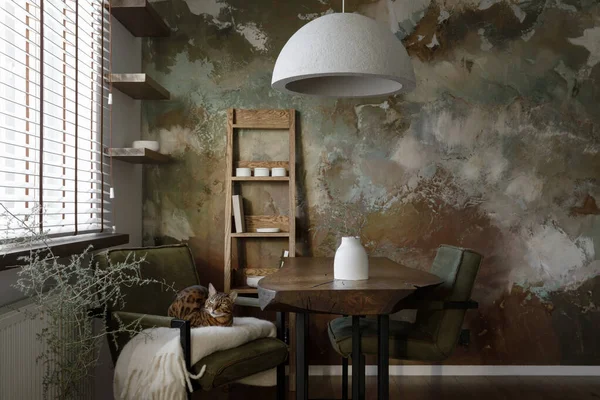 Stylish forest green interior of dining room design with solid oak wooden table, oak shelf and green chairs, bengal cat, concrete vase, elegant cement accessories, recycled paper lamp shade and clay wall in earth tones