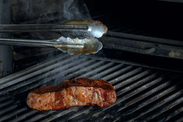 Silver serving kitchen tongs holding raw meat beef steak bbq grilling on rack charcoal stove with smoke on black background.