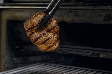 Gümüş servis edilen mutfak maşaları et bifteği ızgara kömür ocağında ızgara ve siyah arka planda duman..