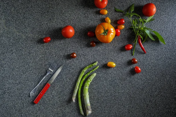Food border with green asparagus, red and orange tomatoes and red chilli pepper with leaves. Vegetables, red knife and fork on dark grey background  top view, place for text, border