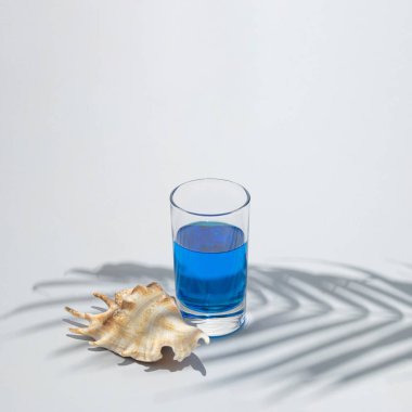 Palmiye dalının altındaki bir bardakta mavi içecekle deniz kabuğu. Palmiye ağacı gölgesi plaj yaz gününü taklit ediyor. Soyut estetik tasarım fikri. Güneşli gün konsepti.