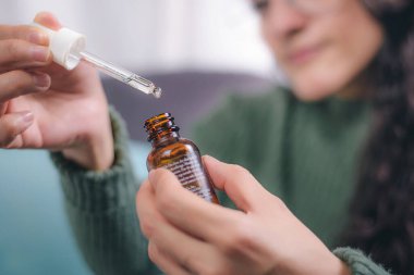 İlgi ve güzellik kavramı. Elinde damlalık olan bir konteynır tutan genç kadının elleri içeriği gösteriyor. Kadın evinde bir koltukta oturuyor..