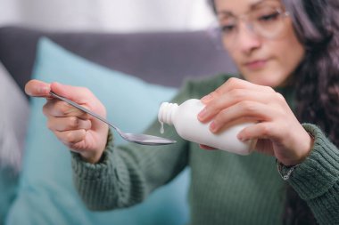 Tıp konsepti. Genç bir kadının elleri kaşıkta öksürük şurubu tutarken, kadın evinde bir koltukta oturuyor..