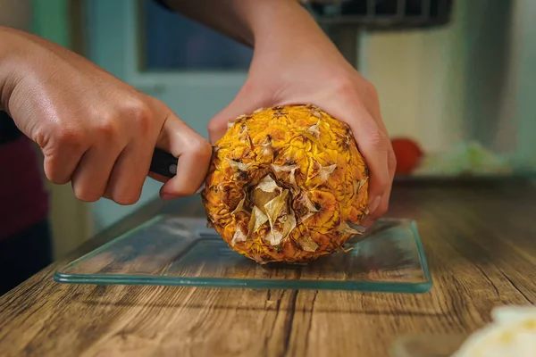 Close-up of a young woman's hands cutting a juicy pineapple with a chef's knife on a wooden table in a kitchen.