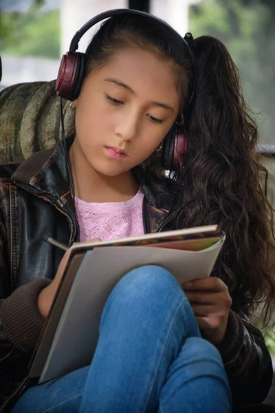 十几岁的女孩坐在公园的台阶上 一边在笔记本上写字 一边带着耳机听音乐 图库照片
