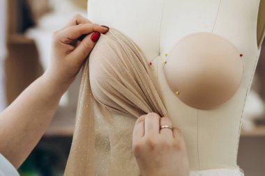 İşteki kadın yeni kumaşını bir mankenin üzerinde sunuyor. Kumaş açık kahverengidir. Mankenin içinde birkaç iğne var..