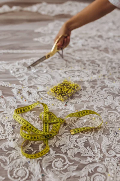 A professional person is cutting the fabric of white ornament with yellow scissors. She has yellow scissors in her right hand. In the front we can see a ruler and a box of yellow needles.