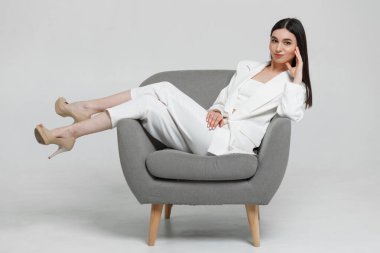 Beyaz takım elbiseli güzel bir kadın rahat gri bir koltukta oturuyor gülümsüyor ve kameraya bakıyor.