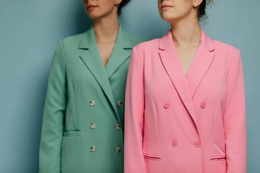 Mavi duvarın önünde soldan bakan renkli takım elbiseli iki güzel kadın manken.