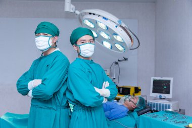 Dortör Profesör, gülümseyen cerrah ameliyathanede dururken kollarını kavuşturuyor..