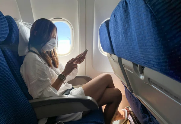 Woman sitting at airplane looking mobile phone and wearing face mask.