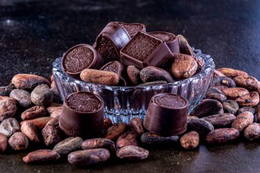 Oyma cam tepside çikolatalar, kakao çekirdeği ile koyu bir arka planda fotoğraflanmış..