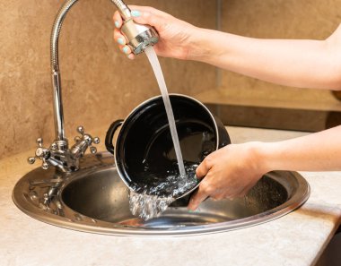 Small metal washbasin with mixer fitted with flexible hose, silver color. female hands hold a container under a stream of water.