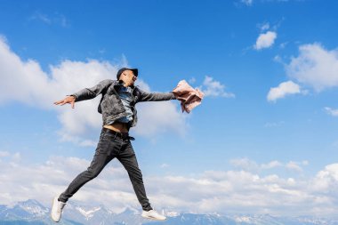 Beyaz bulutlu mavi gökyüzünün arka planına karşı genç bir adam kollarını ve bacaklarını farklı yönlere açarak sıçrıyor..