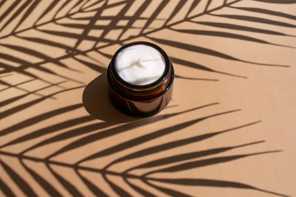 Kosmetyczny krem w ciemnym szklanym słoiku na beżowym tle. Organiczny balsam nawilżający z cieniem liści palmowych. — Zdjęcie stockowe