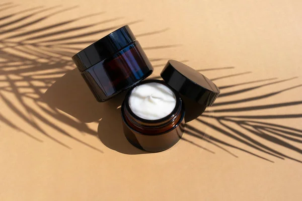 Dwa kremy kosmetyczne w ciemnym szklanym słoiku na beżowym tle. Organiczny balsam nawilżający z cieniem liści palmowych. — Zdjęcie stockowe