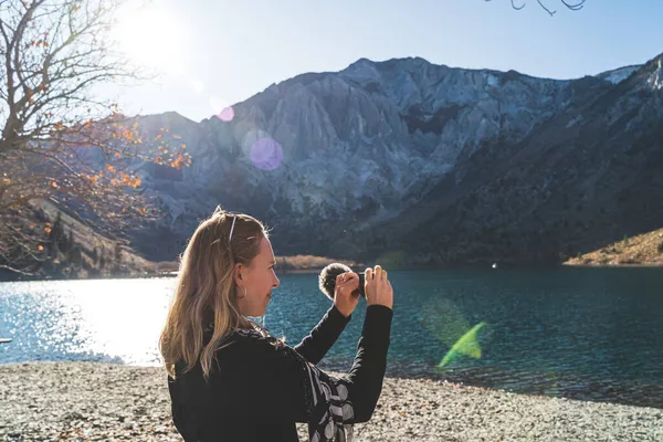 Female travel influencer filming with video camera in front of mountain lake — Stock Photo, Image