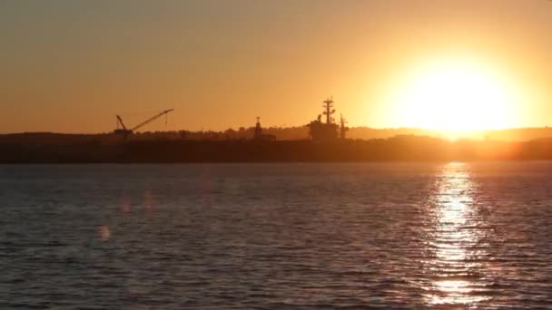 Sun setting over water in bay with silhouette of navy ship and crane — Stock Video