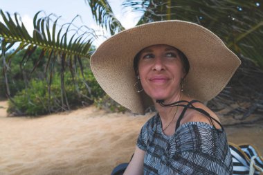 Hasır şapkalı bir kadın tropik plajda oturuyor.