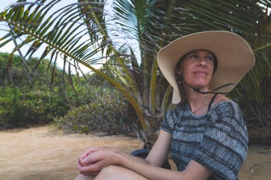 Hasır şapkalı bir kadın tropikal plajda oturmuş, yüzünü yana doğru çeviriyor.