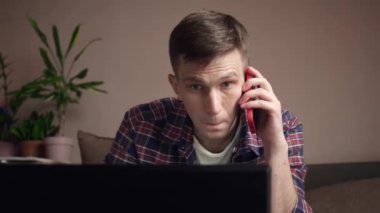 Dizüstü bilgisayar kullanan, koltukta otururken cep telefonuyla konuşan gülümseyen genç adam. Uzaktan çalışma. yakın plan.