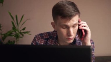 Dizüstü bilgisayar kullanan, koltukta otururken cep telefonuyla konuşan gülümseyen genç adam. Uzaktan çalışma. yakın plan