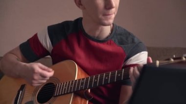 Genç adam evde online olarak akustik gitar çalmayı öğreniyor. yakın plan.